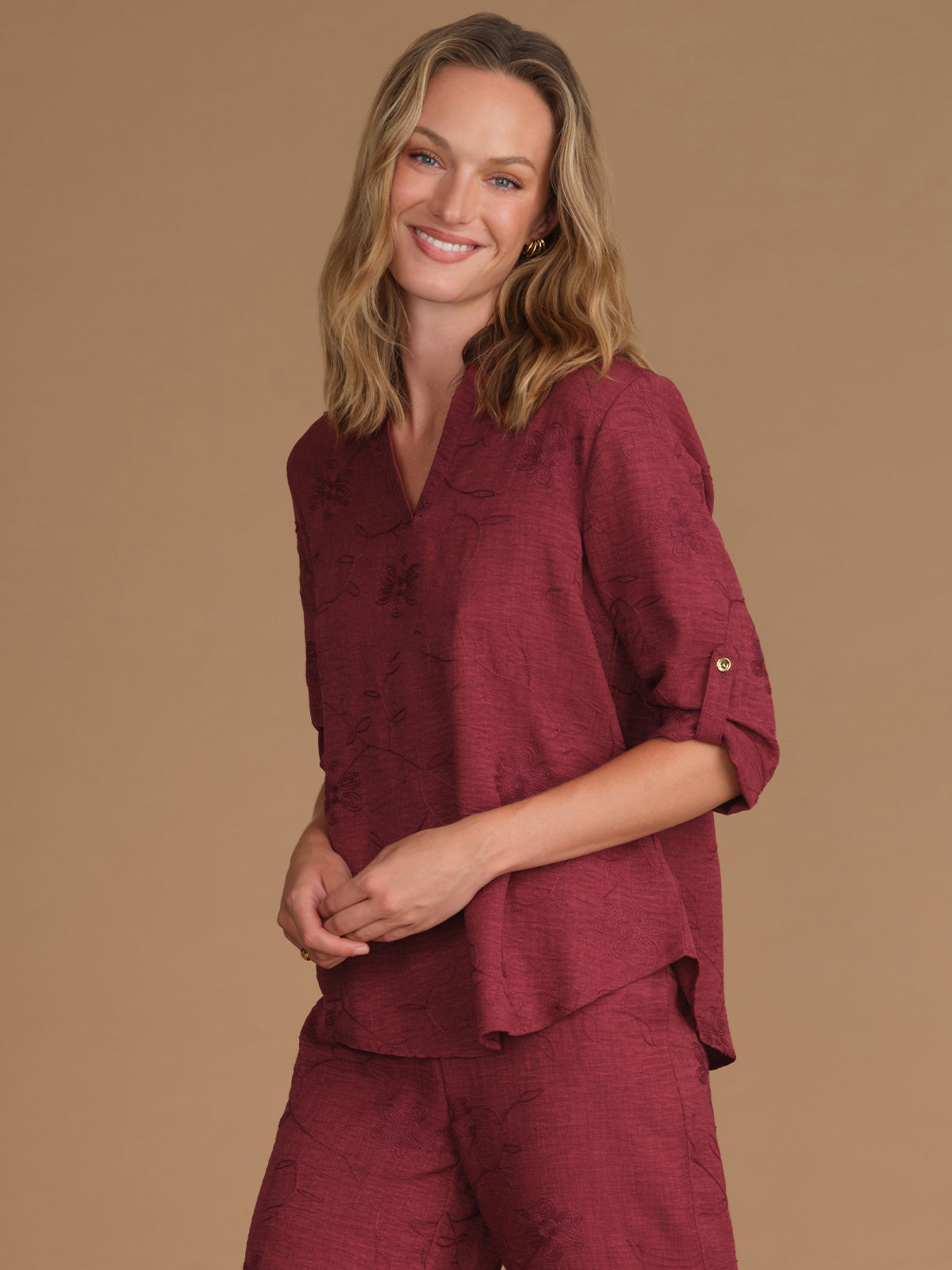 A woman modeling a matching burgundy tops with a subtle tonal floral jacquard pattern, featuring a short-sleeve top with rolled cuffs and matching pants, set against a solid tan studio background.