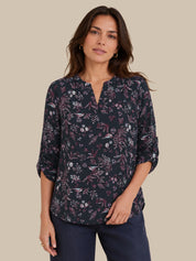 A woman modeling a dark navy floral blouse with a split neckline and three-quarter sleeves, paired with dark blue jeans, against a solid beige background.