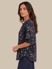 A woman wearing a dark navy floral and bird-patterned top with three-quarter sleeves, shown in a side-profile view against a solid beige background.