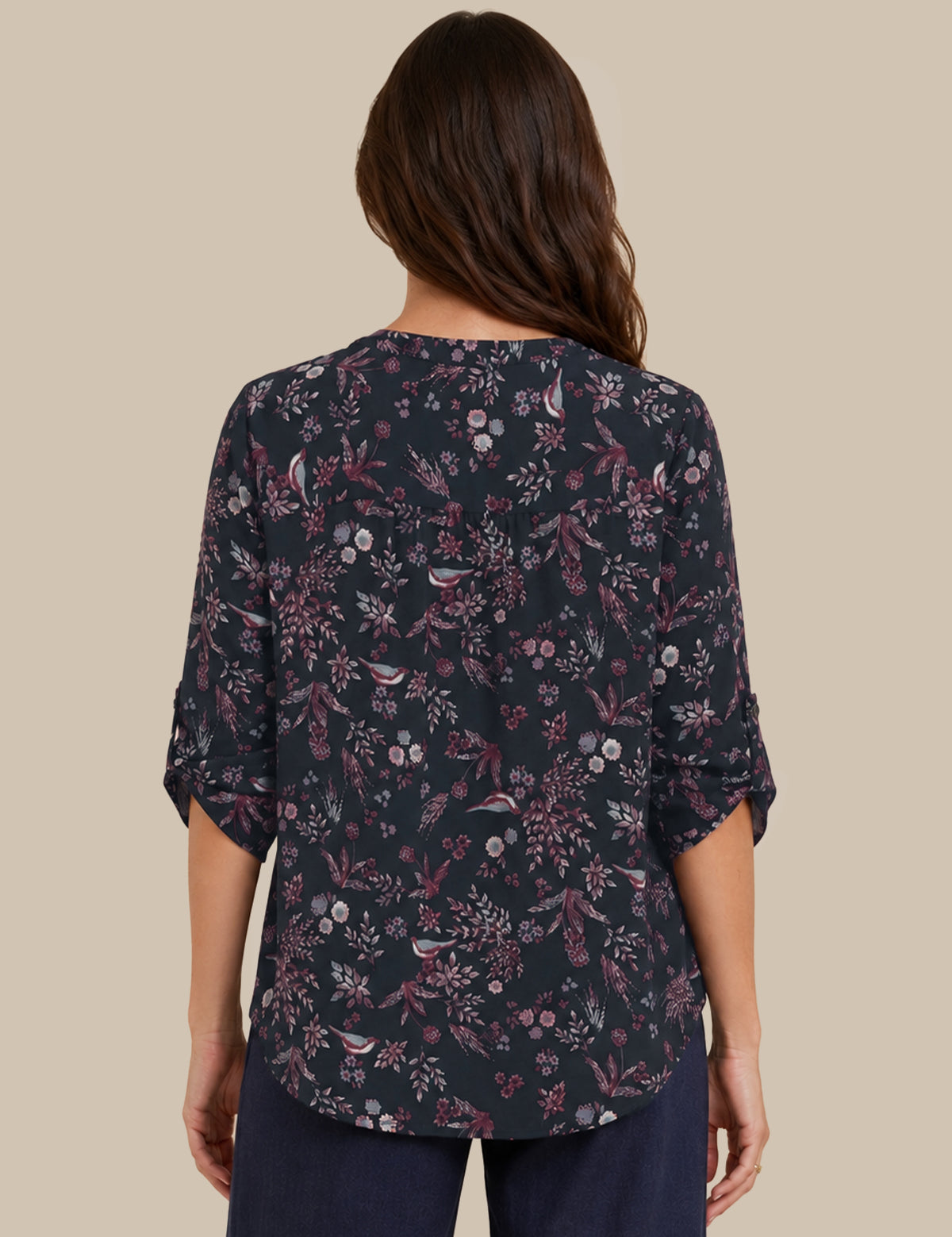 A woman viewed from the back wearing a dark navy floral blouse with three-quarter sleeves and dark navy pants, set against a solid beige background.