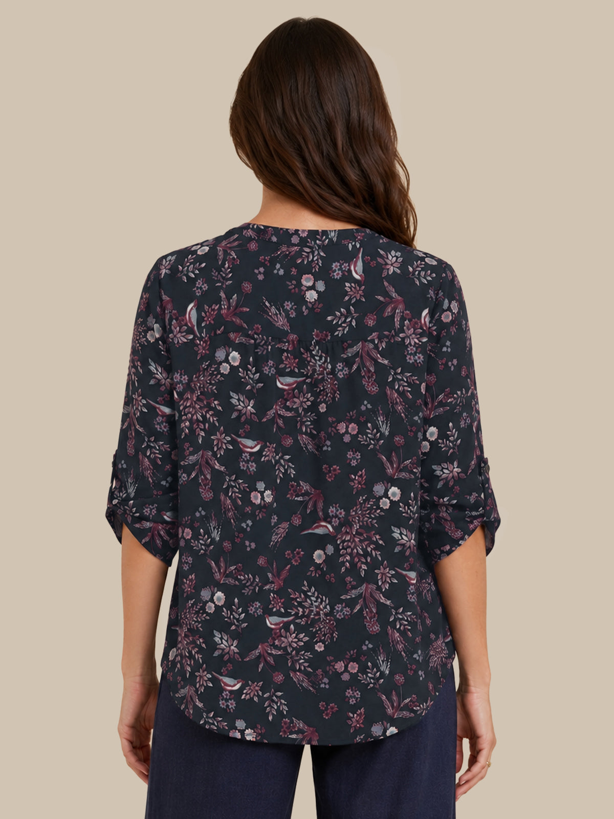 A woman viewed from the back wearing a dark navy floral blouse with three-quarter sleeves and dark navy pants, set against a solid beige background.