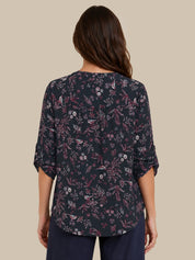 A woman viewed from the back wearing a dark navy floral blouse with three-quarter sleeves and dark navy pants, set against a solid beige background.
