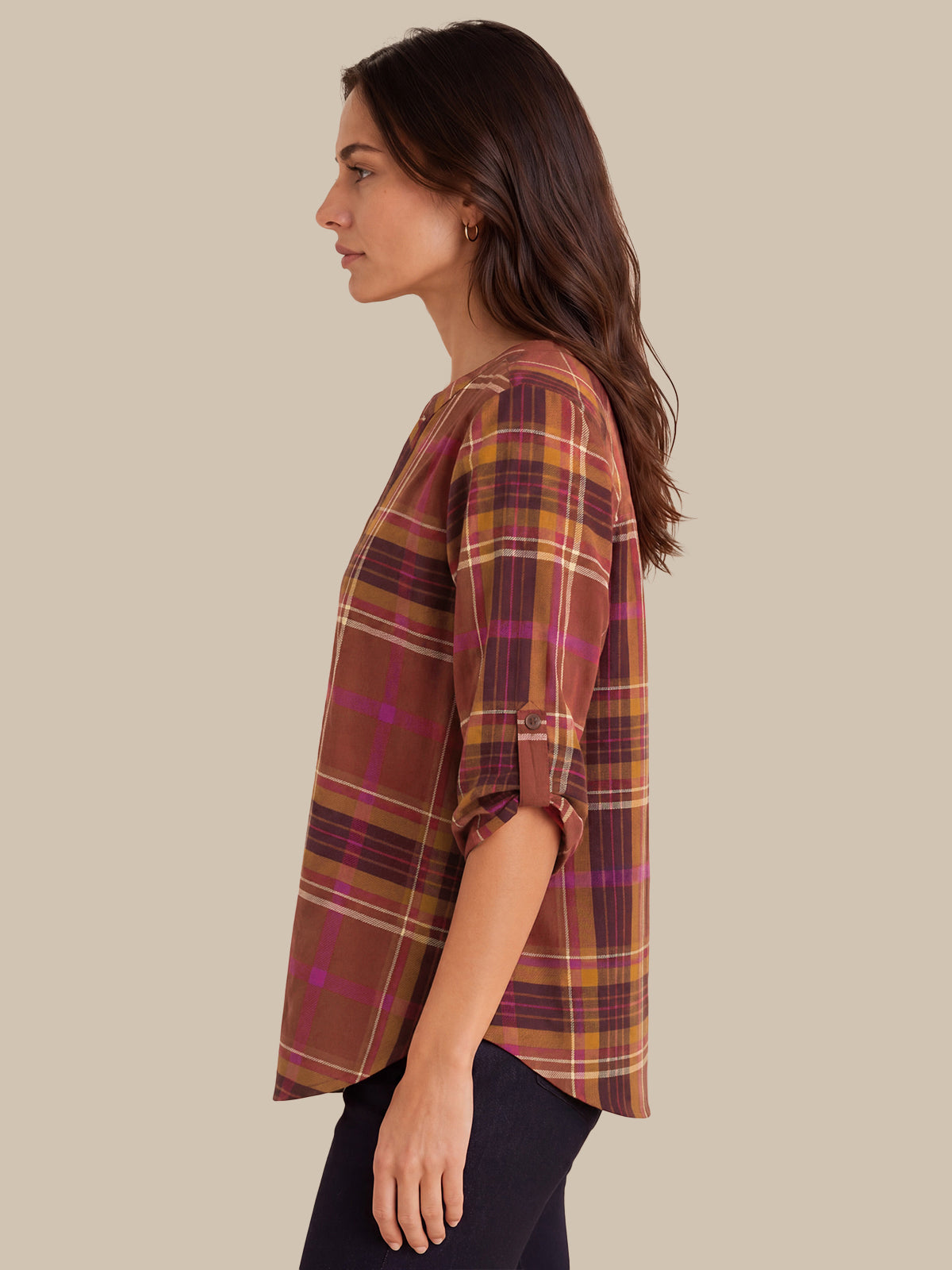 A woman shown in side profile wearing a loose-fitting plaid top in rust brown, mustard yellow, and magenta tones, paired with dark pants, against a solid beige background.