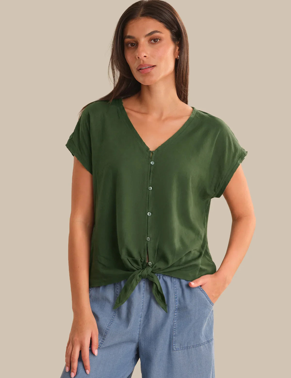 Woman wearing a green top with a tie detail and blue pants on a beige background