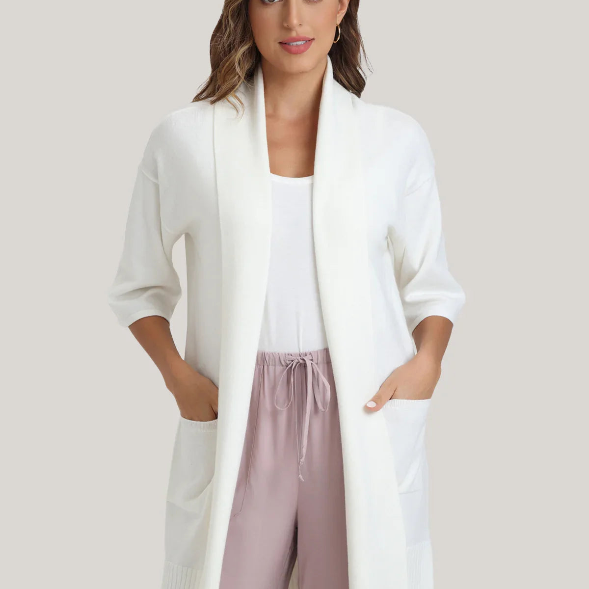 Woman wearing a white cardigan over a white top and pink pants on a light background