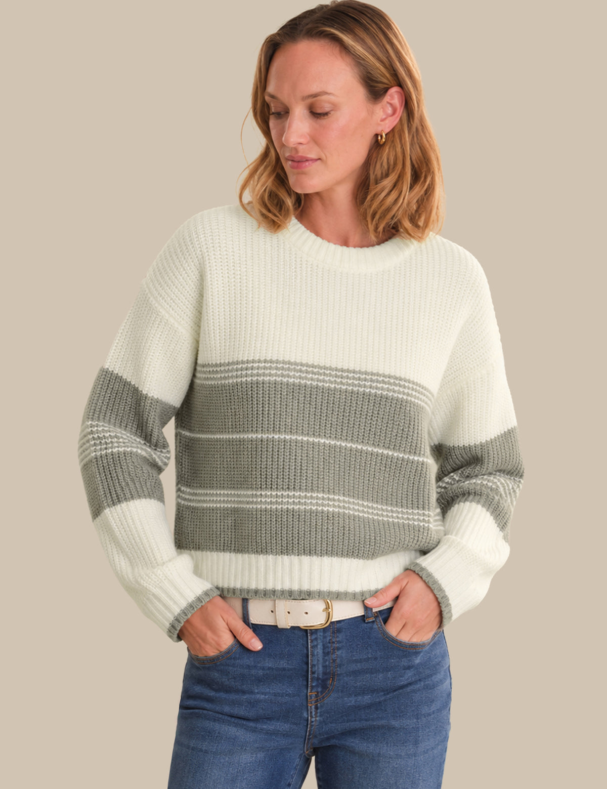 A woman wearing a ribbed knit sweater with horizontal stripes in off-white and sage green, paired with blue jeans, against a solid beige background.