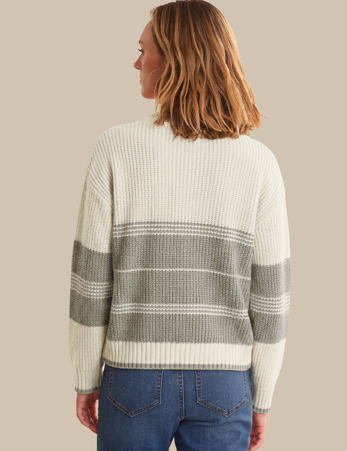 A woman shown from the back wearing a ribbed knit sweater with horizontal stripes in cream and gray, paired with blue jeans, against a solid beige background.