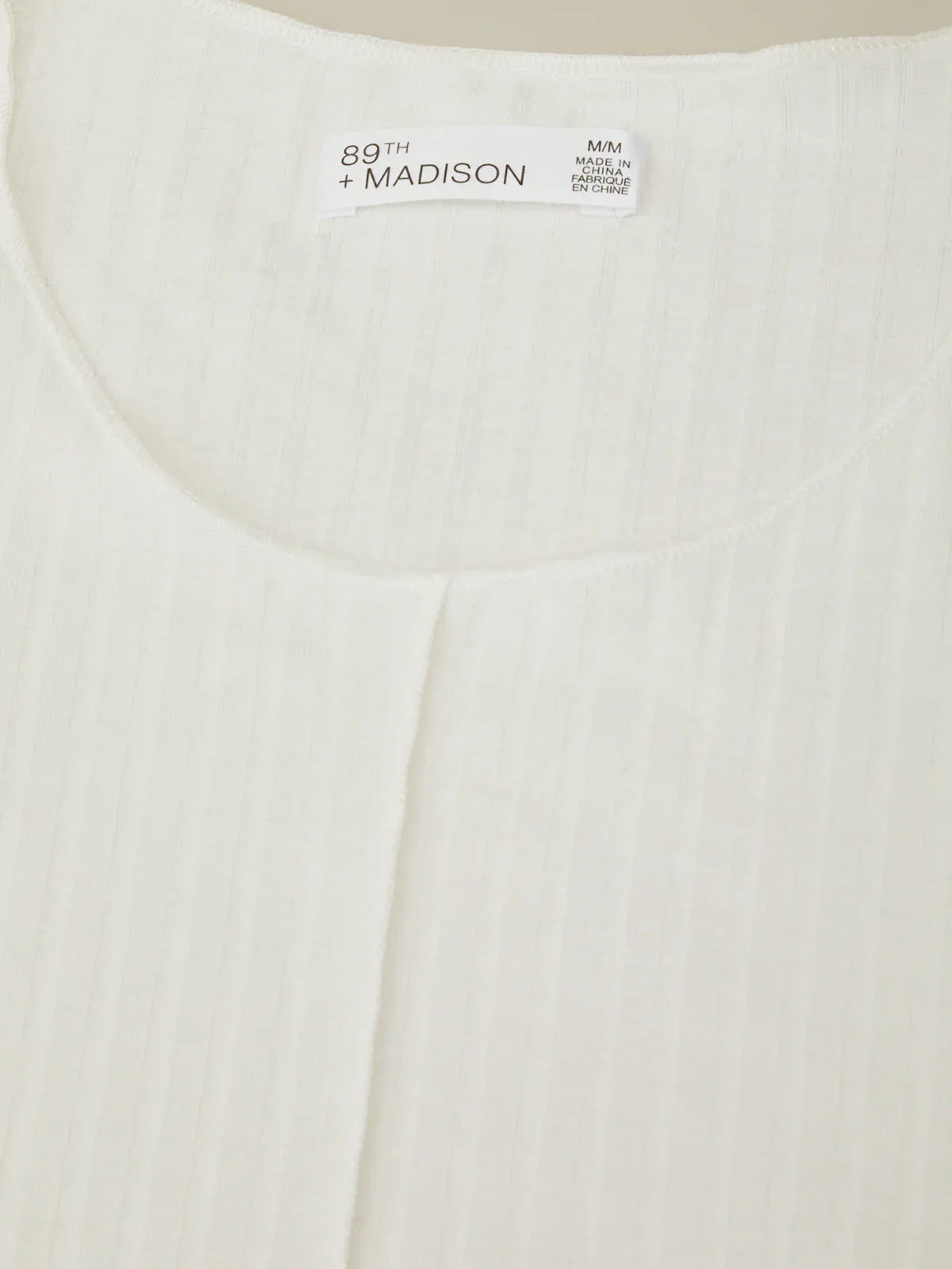 Close-up of a white garment with '89th + Madison' branding on a beige background