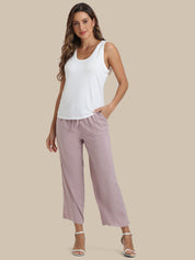A woman modeling a white sleeveless tank top and dusty lavender cropped drawstring pants against a solid beige background.