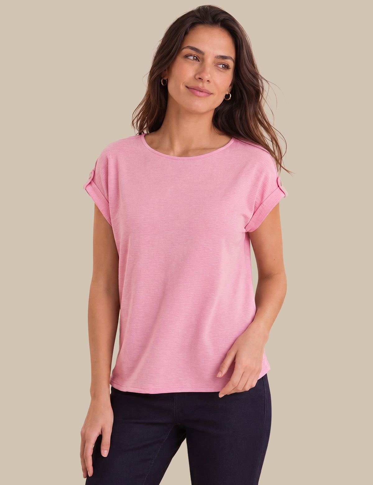 A woman modeling a light pink short-sleeve top with rolled cuffs, paired with dark navy pants, against a solid beige background.