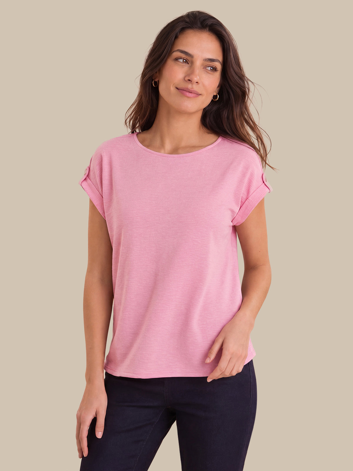 A woman modeling a light pink short-sleeve top with rolled cuffs, paired with dark navy pants, against a solid beige background.