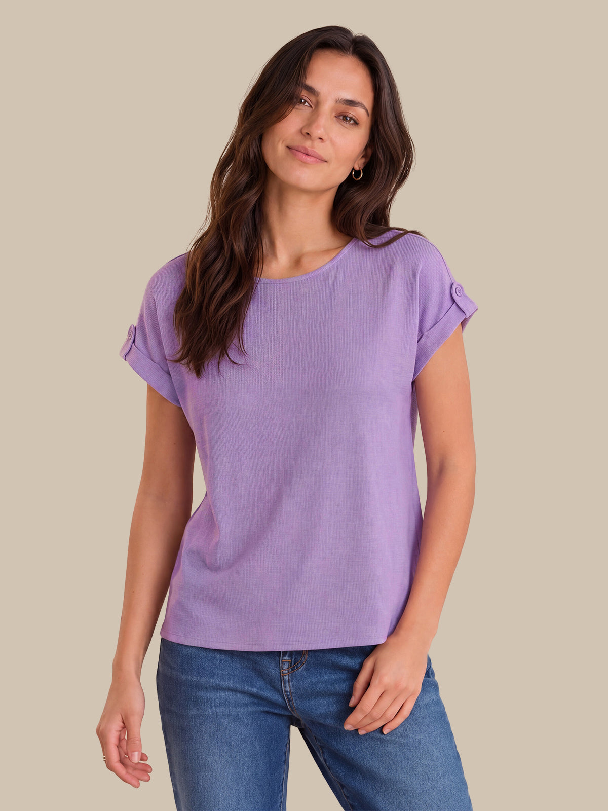 A woman modeling a lavender purple short-sleeve top with rolled cuffs, paired with blue jeans, against a solid beige background.