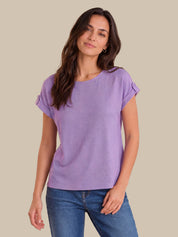 A woman modeling a lavender purple short-sleeve top with rolled cuffs, paired with blue jeans, against a solid beige background.