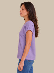 A woman shown in side profile wearing a light purple short-sleeve knit top and blue jeans against a solid beige background.