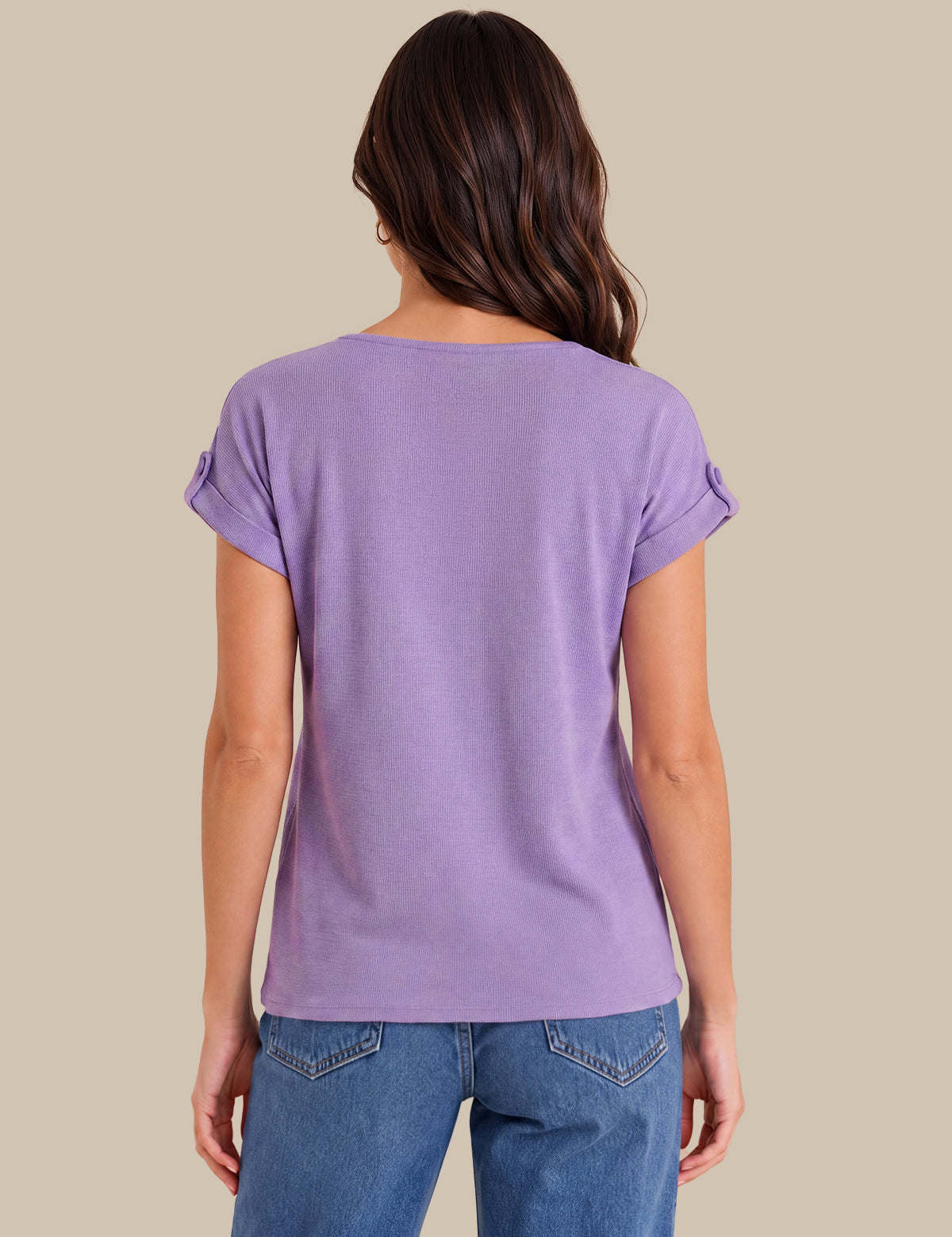 A woman viewed from the back wearing a lavender purple short-sleeve t-shirt with rolled cuffs and blue jeans, set against a solid beige background.