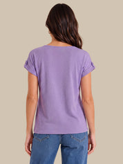 A woman viewed from the back wearing a lavender purple short-sleeve t-shirt with rolled cuffs and blue jeans, set against a solid beige background.