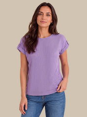 A woman modeling a lavender short-sleeve crewneck t-shirt with rolled cuffs, paired with blue jeans, against a solid beige background.