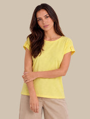 A woman modeling a bright yellow short-sleeve t-shirt with ruffled cuffs, paired with beige pants, against a solid beige background.