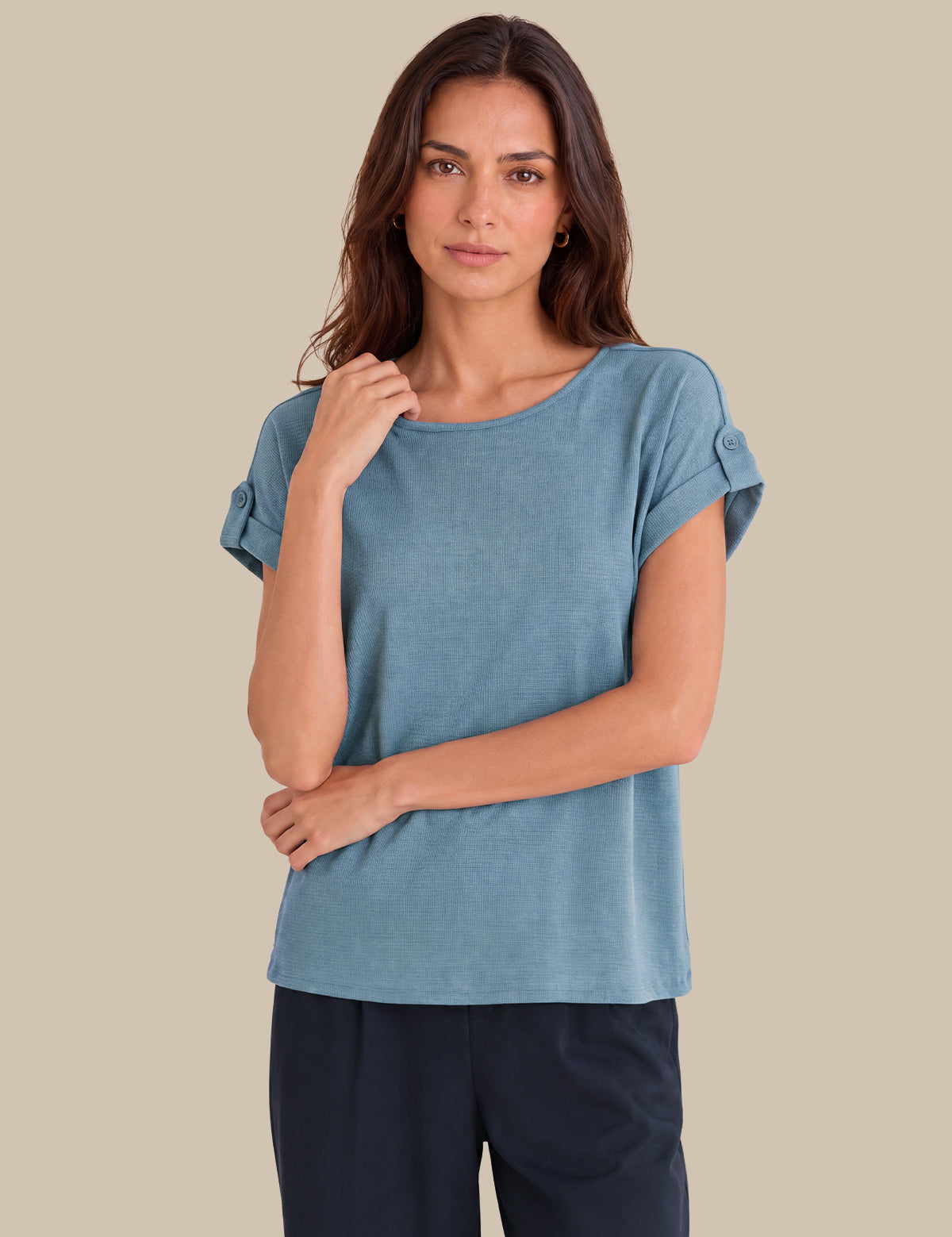 A woman modeling a dusty blue short-sleeve top with rolled cuffs, paired with dark navy pants, against a solid beige background.