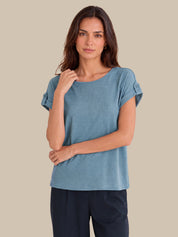 A woman modeling a dusty blue short-sleeve top with rolled cuffs, paired with dark navy pants, against a solid beige background.
