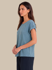 A woman wearing a dusty blue short-sleeve top with rolled cuffs and dark navy pants, standing against a solid beige background.
