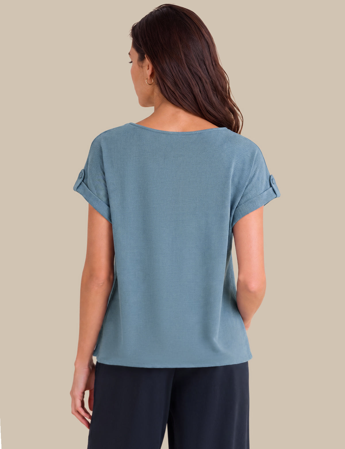 A woman shown from the back wearing a dusty blue short-sleeve top with rolled cuffs and dark navy pants, set against a solid beige background.