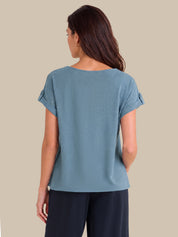 A woman shown from the back wearing a dusty blue short-sleeve top with rolled cuffs and dark navy pants, set against a solid beige background.