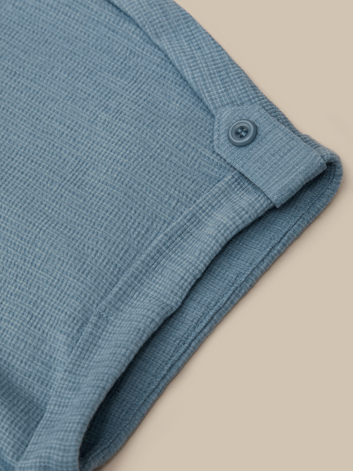 Close-up of a dusty blue textured knit garment, showing a folded cuff with a matching button, set against a solid beige background.