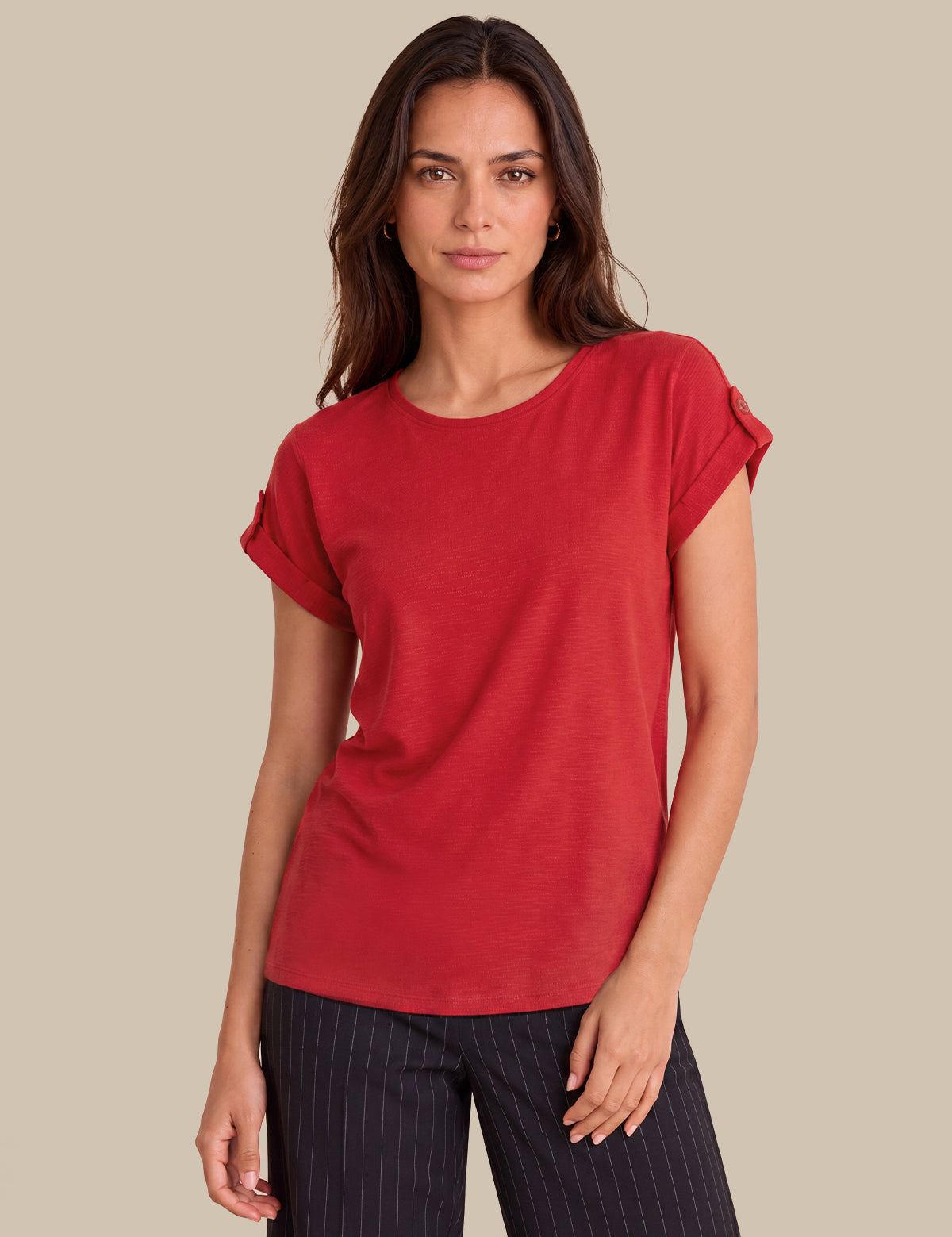 A woman modeling a red short-sleeve top with rolled cuffs, paired with dark pinstriped pants, against a solid beige background.
