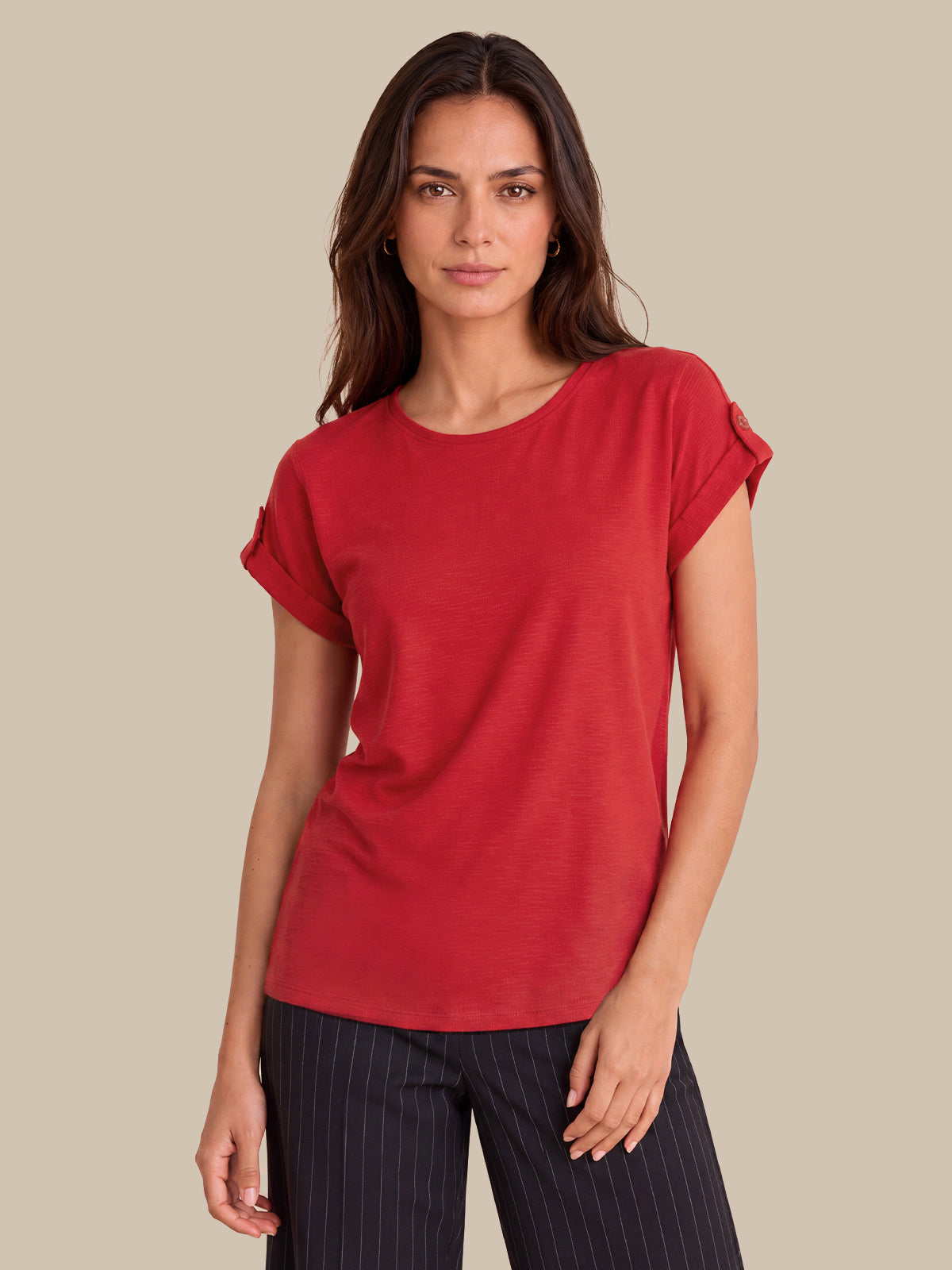 A woman modeling a red short-sleeve top with rolled cuffs, paired with dark pinstriped pants, against a solid beige background.