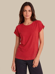 A woman modeling a red short-sleeve top with rolled cuffs, paired with dark pinstriped pants, against a solid beige background.