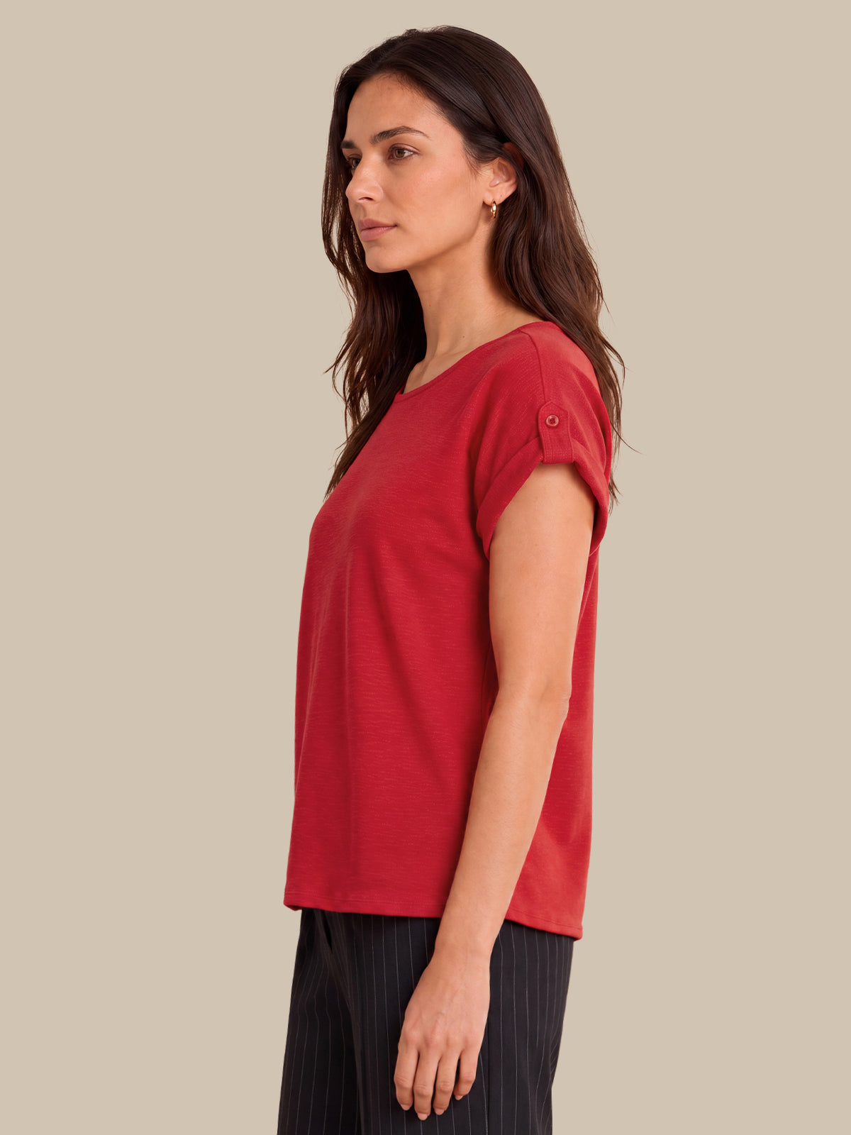 A woman shown in side profile wearing a red short-sleeve top with rolled cuffs and dark pleated trousers, against a plain beige studio background.
