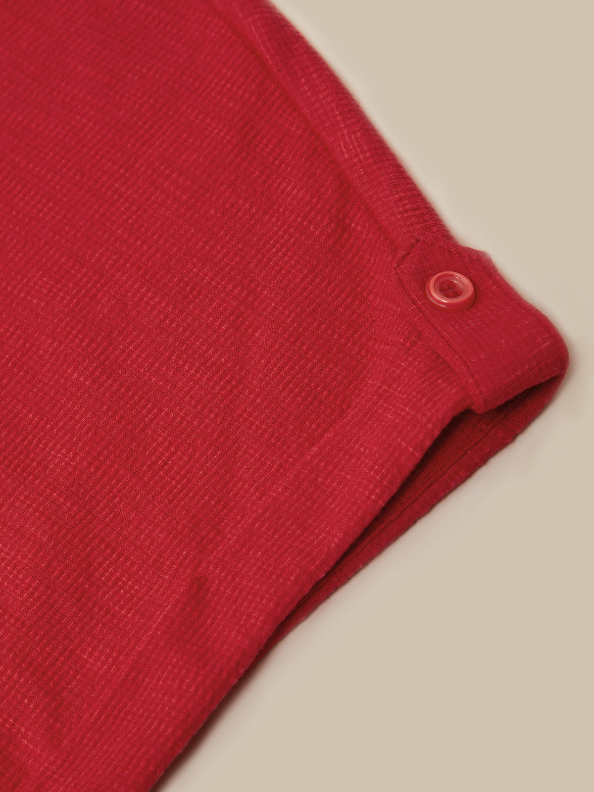 Close-up of a folded red knit garment, showing the fabric texture and a small matching red button tab near the cuff, on a light beige background.