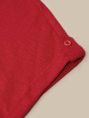Close-up of a folded red knit garment, showing the fabric texture and a small matching red button tab near the cuff, on a light beige background.