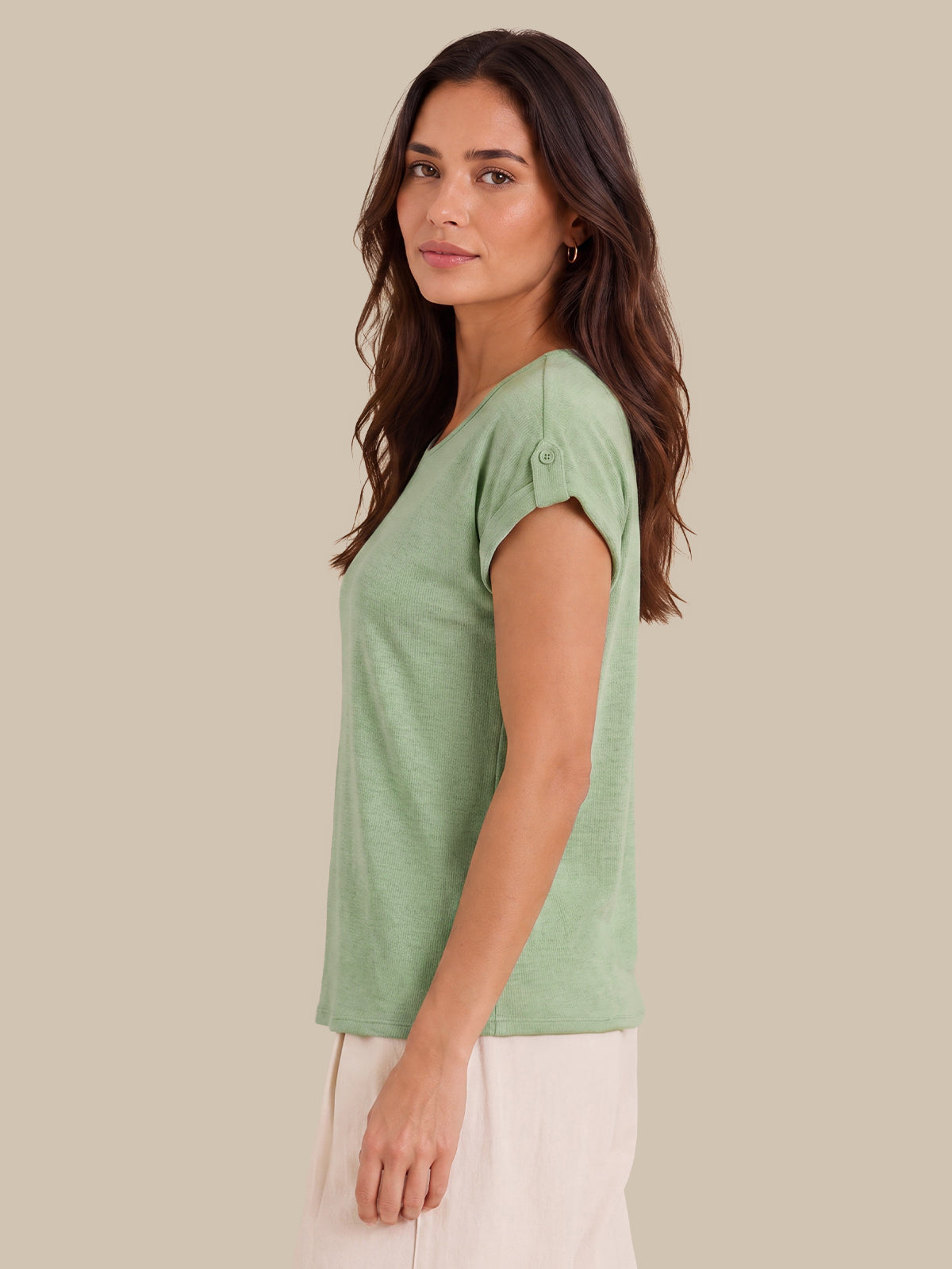 A woman wearing a light sage green short-sleeve top with button details on the sleeve cuffs, paired with off-white pants, shown in profile against a solid beige background.