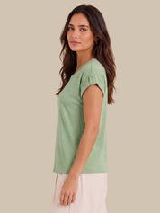 A woman wearing a light sage green short-sleeve top with button details on the sleeve cuffs, paired with off-white pants, shown in profile against a solid beige background.