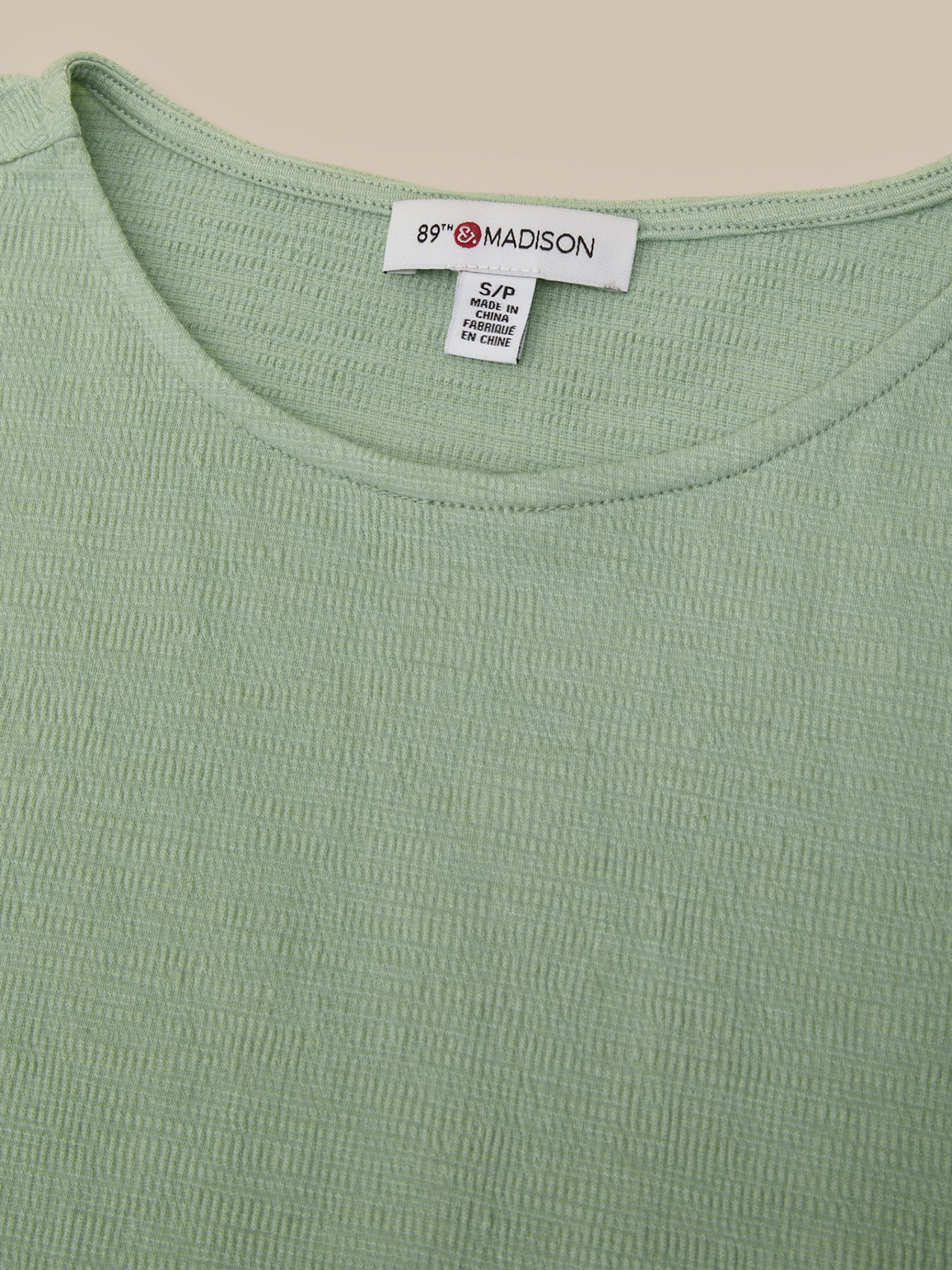 Close-up of a light sage green ribbed knit top with a white neck label reading 89° MADISON.