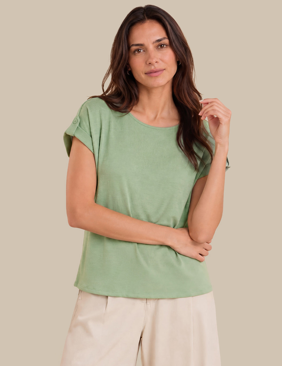 A woman modeling a sage green short-sleeve top with rolled cuffs and light beige pants, set against a solid beige background.