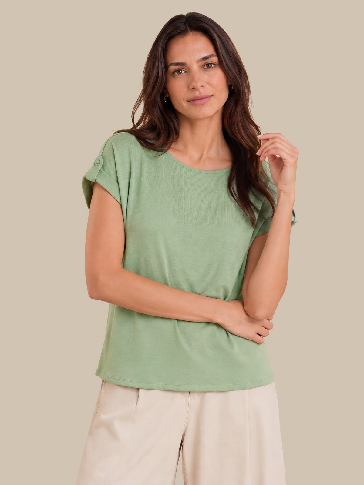 A woman modeling a sage green short-sleeve top with rolled cuffs and light beige pants, set against a solid beige background.