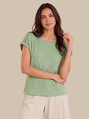 A woman modeling a sage green short-sleeve top with rolled cuffs and light beige pants, set against a solid beige background.
