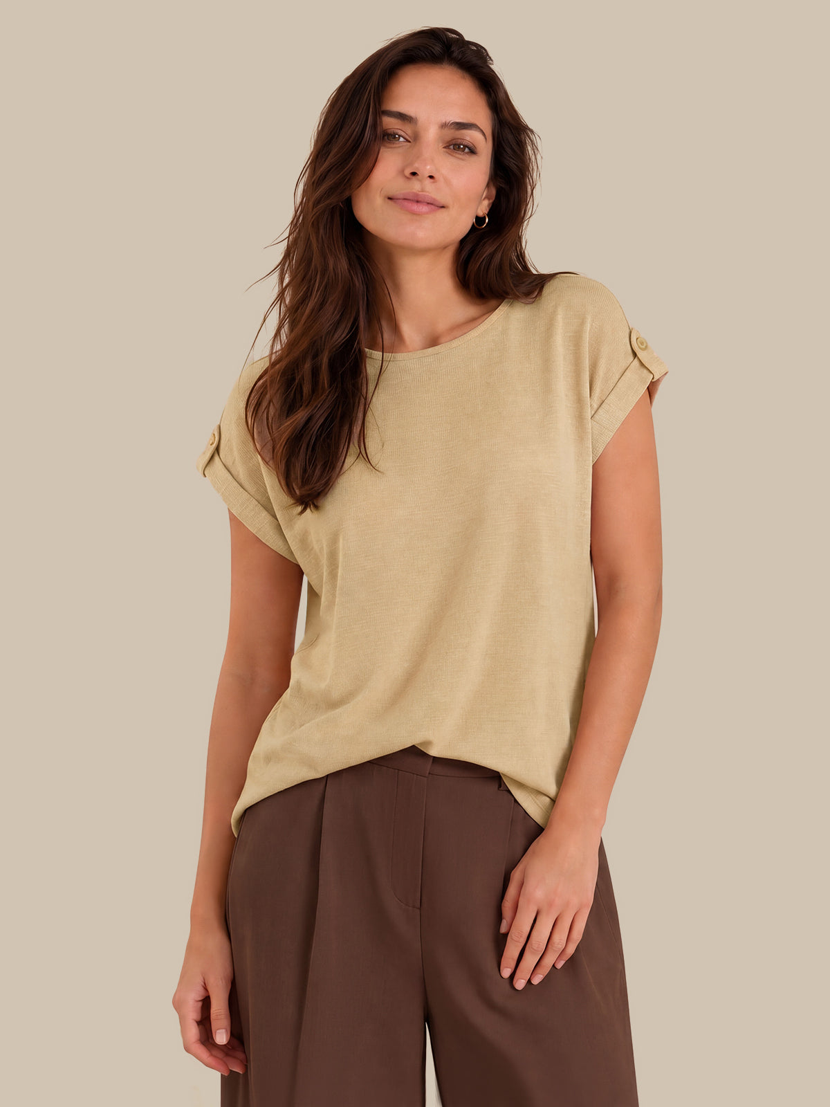 A woman modeling a light beige short-sleeve top with rolled cuffs and brown wide-leg trousers, set against a solid beige studio background.