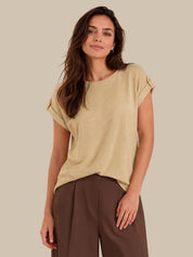 A woman modeling a light beige short-sleeve top with rolled cuffs and brown wide-leg trousers, set against a solid beige studio background.