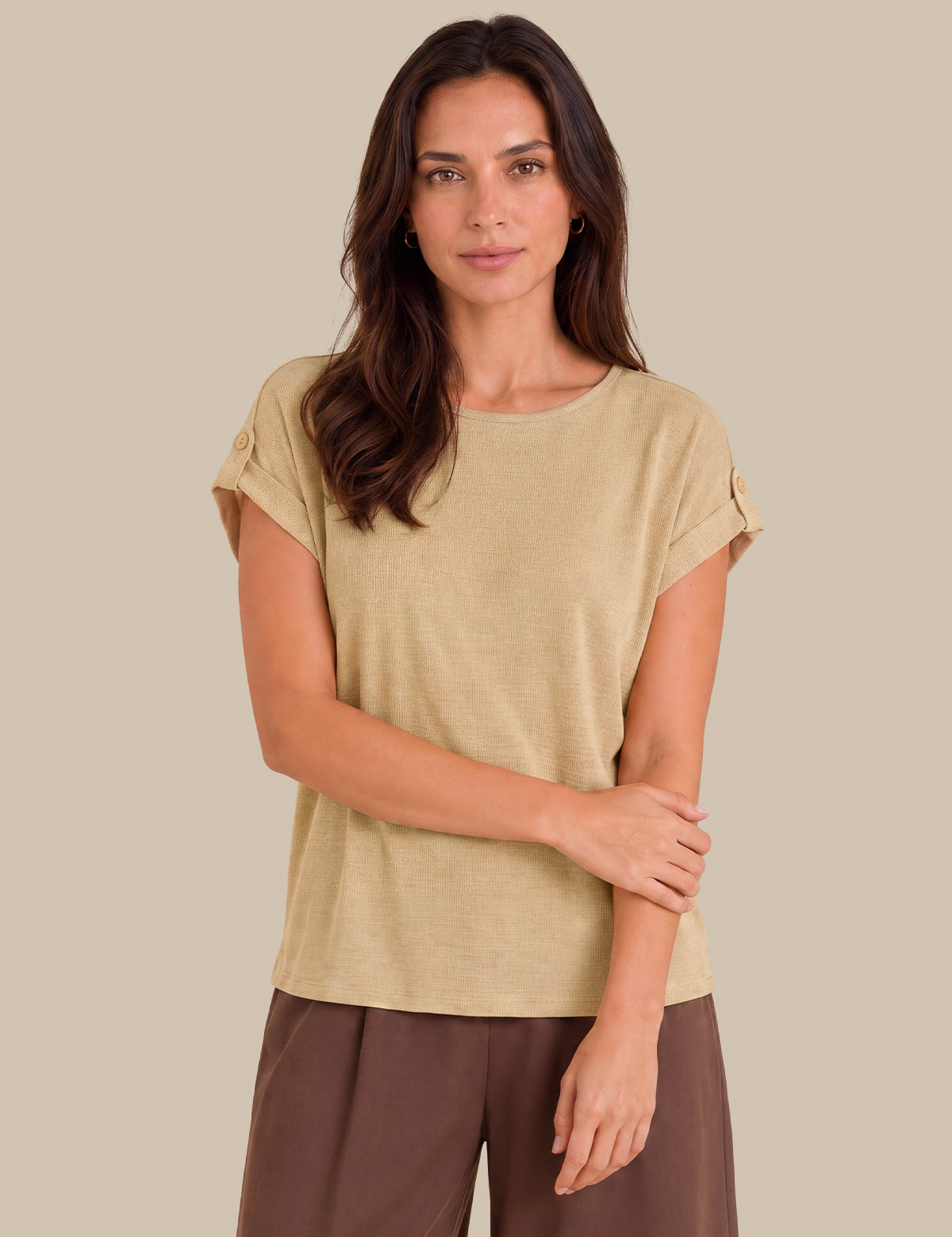 A woman modeling a beige short-sleeve top with rolled cuffs and brown pants against a solid beige background.