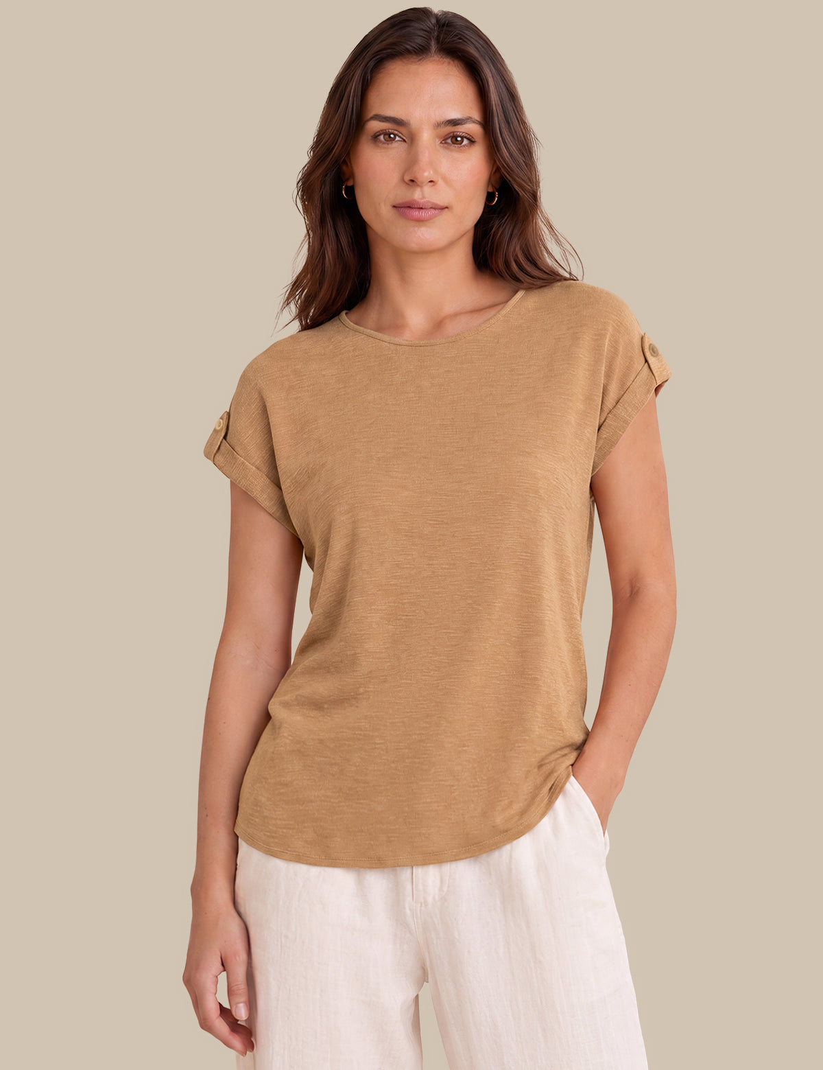 A woman modeling a tan short-sleeve top with rolled cuffs and white pants against a solid beige background.