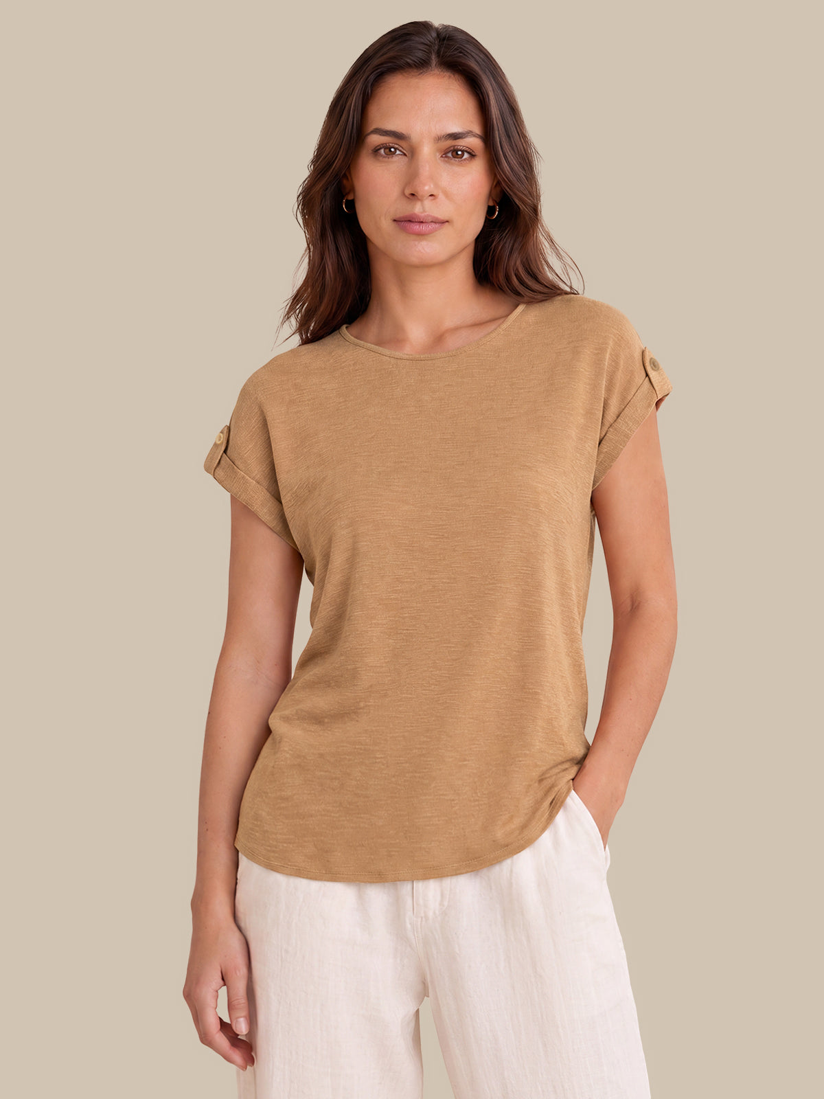A woman modeling a tan short-sleeve top with rolled cuffs and white pants against a solid beige background.