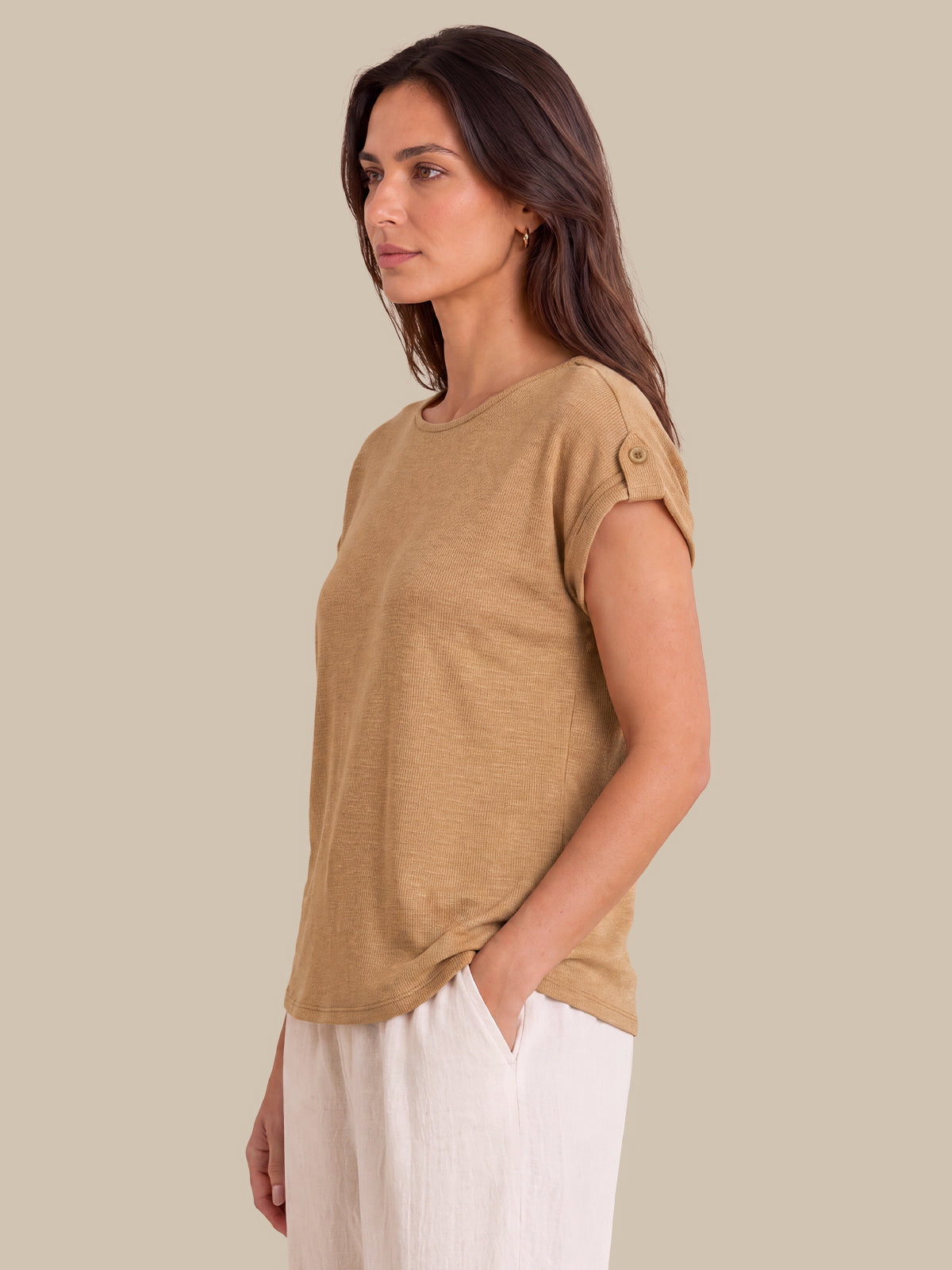 A woman wearing a tan short-sleeve knit top with button details on the sleeve, paired with white pants, shown in profile against a solid beige background.