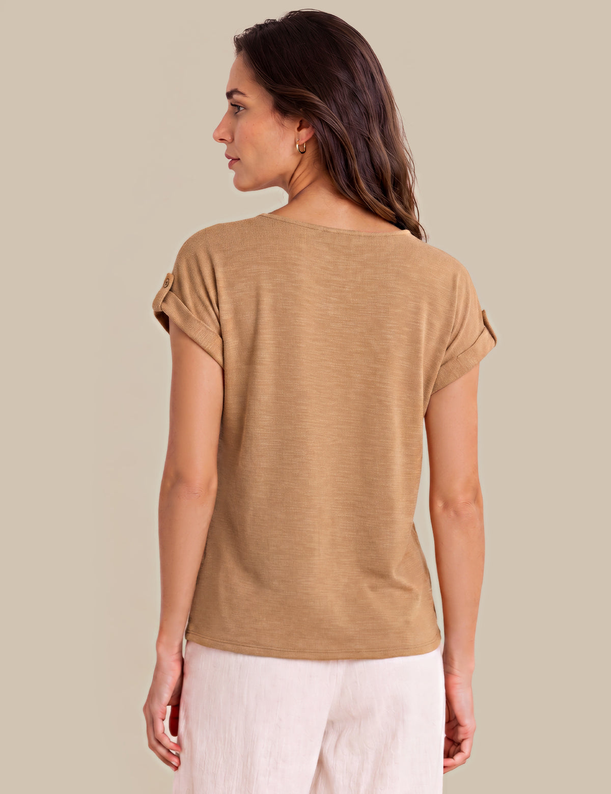 A woman shown from the back wearing a tan short-sleeve top with rolled cuffs and light-colored pants, set against a solid beige background.