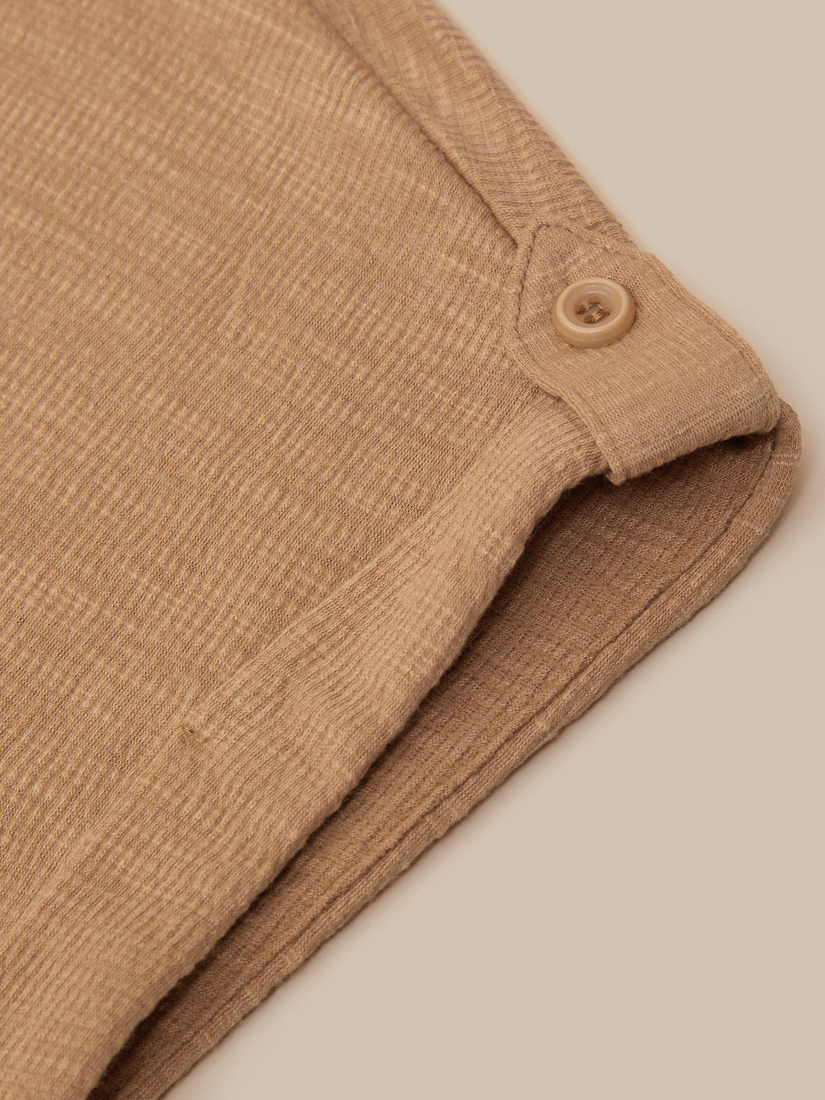 Close-up of a folded tan ribbed fabric item with a small matching button detail, shown on a plain warm beige background.
