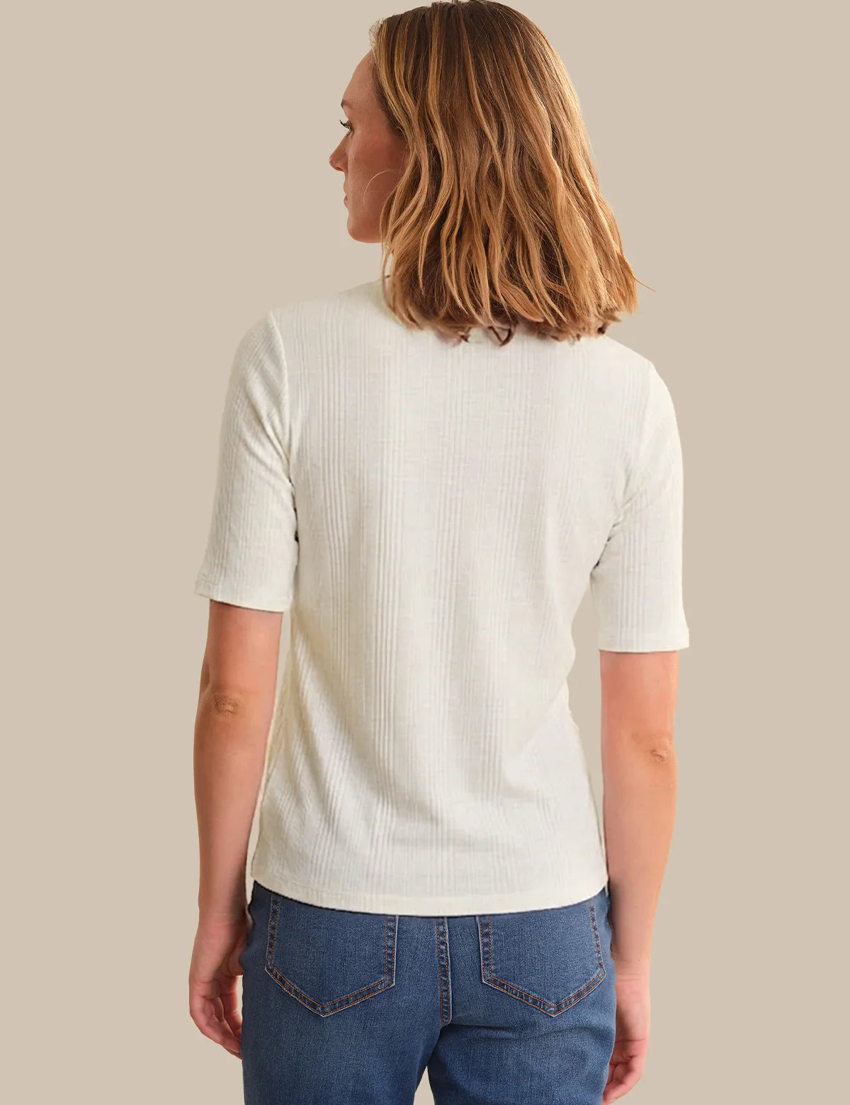 Woman wearing a white ribbed sweater and blue jeans against a beige background