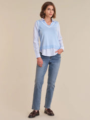 Woman wearing a light blue sweater over a striped shirt with jeans on a beige background